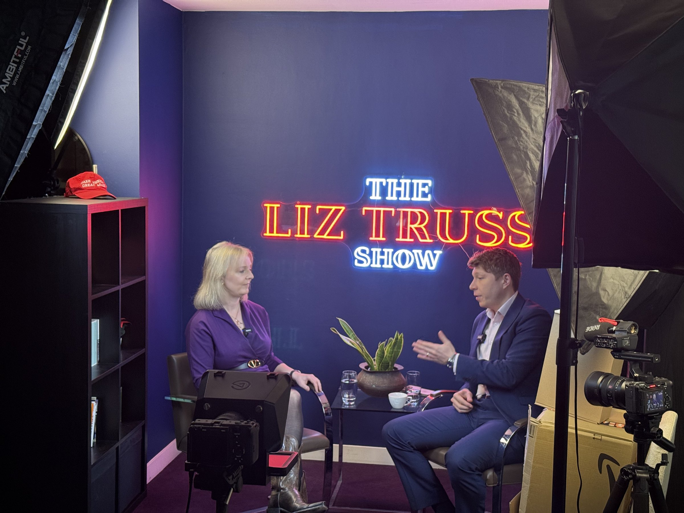 Liz Truss seated on set opposite an interviewer during production of The Liz Truss Show, with the illuminated show sign behind them and cameras, softbox lighting and studio equipment in the foreground.
