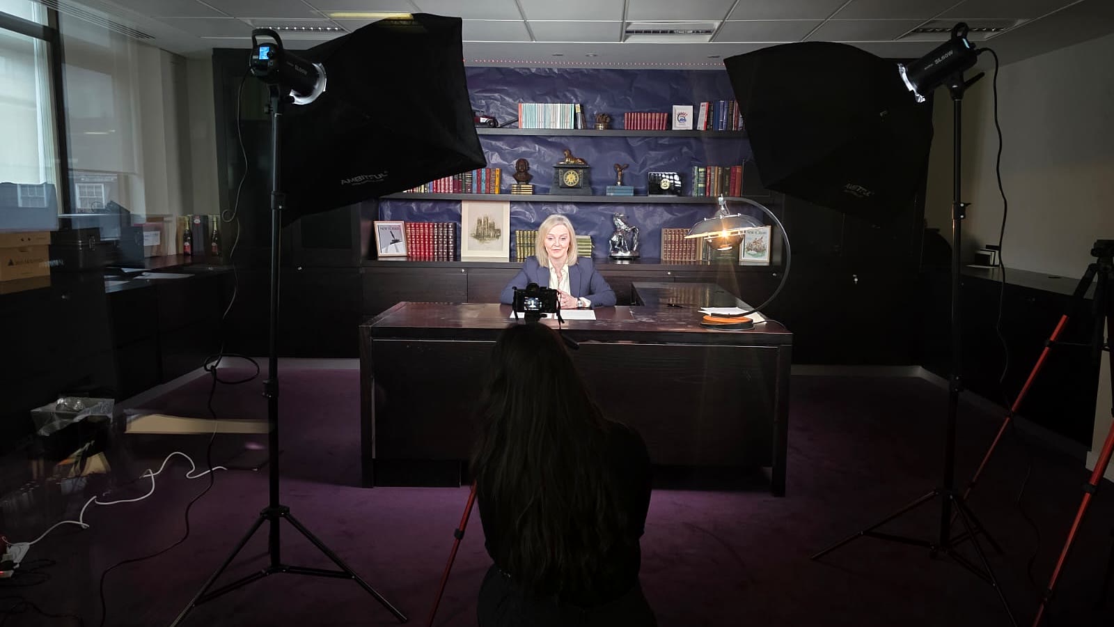 Behind-the-scenes production setup for The Liz Truss Show, with Liz Truss seated at the studio desk between two large softbox lights, while cameras and a crew member are visible in the foreground.